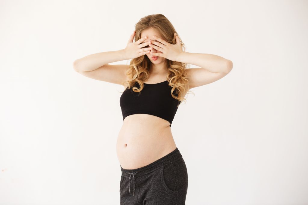portrait-of-funny-european-mother-with-blond-curly-hair-in-black-sport-suit-with-opened-pregnant-belly-clothing-eyes-with-hands-having-surprised-expression.jpg